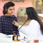 Indian female doctor checking young adult girl patient heartbeat with stethoscope at village outdoor, woman getting examine by medical person, Rural India healthcare concept
